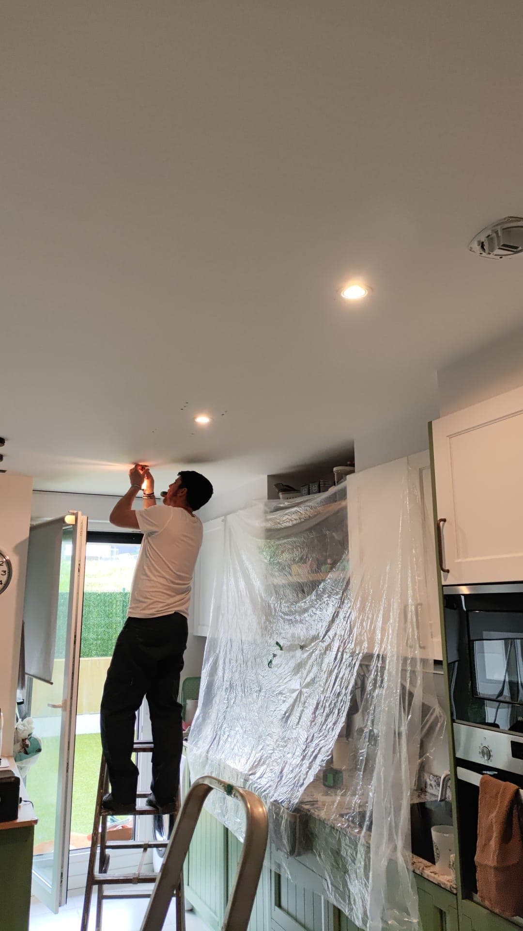 Antonio, subido a una escalera, instalando focos en el techo de una cocina con los muebles tapados con plástico